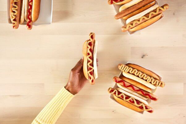 a hand holding the IKEA plant based hot dog on a table surrounded by more with various toppings