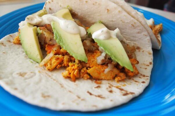 flour tortillas on a blue plate filled with buffalo tofu, potatoes, avocado slices, and topped with vegan ranch dressing