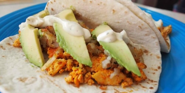 flour tortillas on a blue plate filled with buffalo tofu, potatoes, avocado slices, and topped with vegan ranch dressing