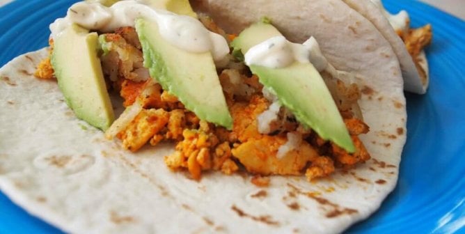 flour tortillas on a blue plate filled with buffalo tofu, potatoes, avocado slices, and topped with vegan ranch dressing