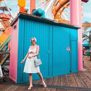 woman in white stands in front of a blue and pink panted building at a carnival