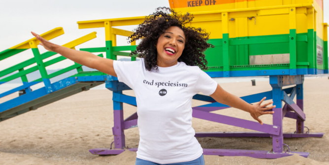 woman with tightly-curled hair jumps on the beach in front of a rainbow painted lifeguard station