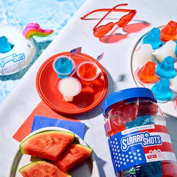 red white and blue vegan gelatin shots from slrrrp shots next to a plate of watermelon, heart-shaped sunglasses, and a pool with a floatie in it