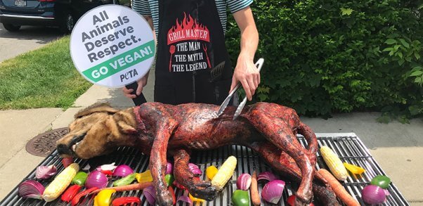 man "grills" a dog prop with a sign in the background that says "all animals deserve respect. go vegan!"
