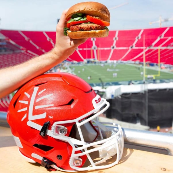 hand holding a vegan lightlife veggie burger, resting on a red football helmet with a stadium in the background
