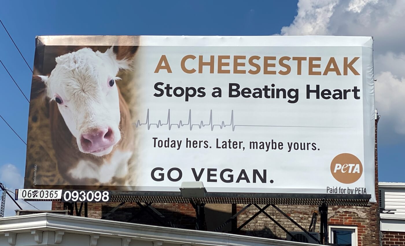 A billboard in Philadelphia, PA featuring a cow reads "A cheesesteak stops a beating heart. Today hers. Later, maybe yours. Go vegan."