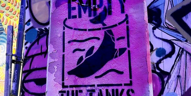 stenciled spray paint art on a concrete pole reading "empty the tanks" with an illustration of a dolphin in a small tank.