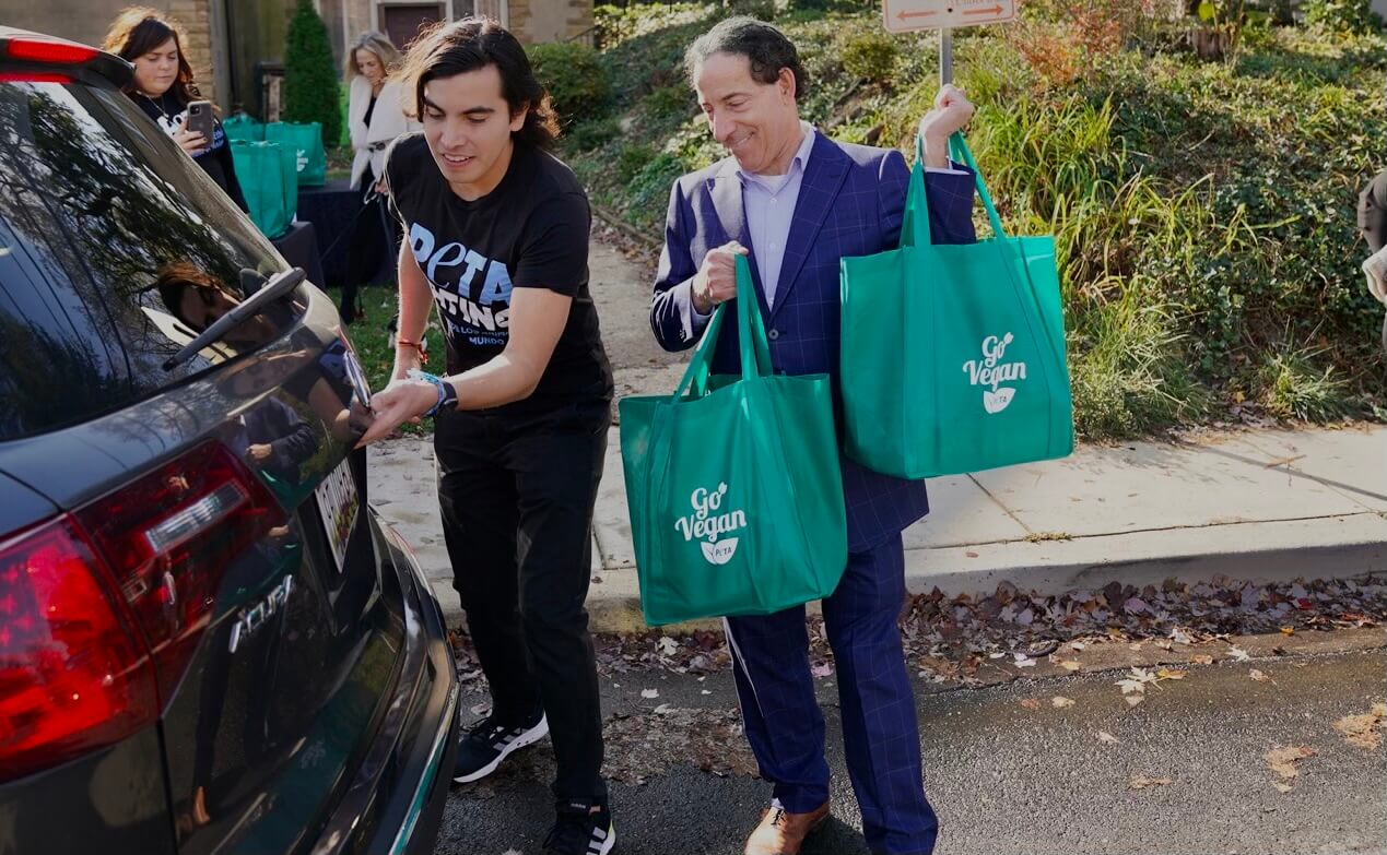 Jamie Raskin lifting two bags toward the trunk of a car, while a person in a PETA shirt opens the trunk