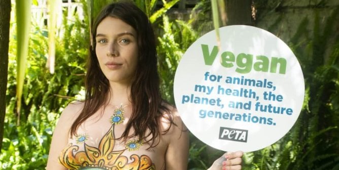 Pregnant protester with painted globe on stomach holding go vegan sign surrounded by plants