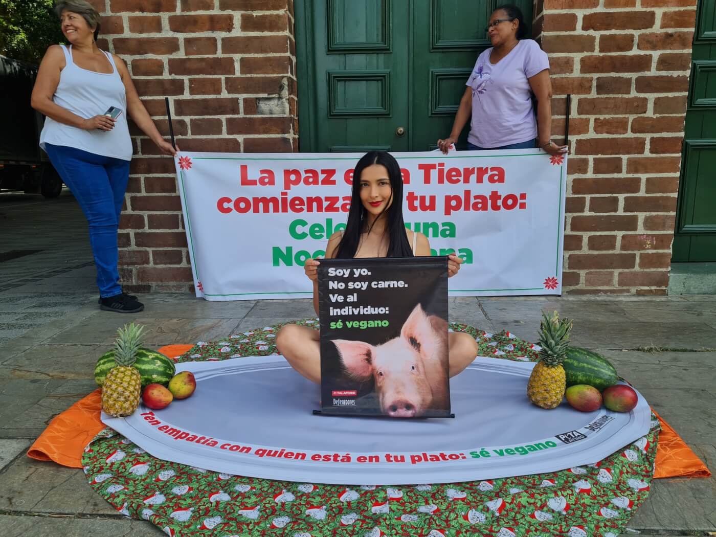 Colombian model Kathy Moscoso sitting on a fake plate surrounded by fruits. She is holding a sign with a pig on it.