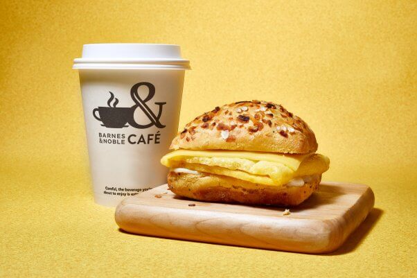 promotional photo of the new vegan breakfast sandwich from barnes & noble on top of a wooden cutting board next to a coffee cup with the barnes & noble cafe logo