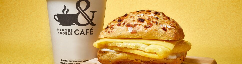 promotional photo of the new vegan breakfast sandwich from barnes & noble on top of a wooden cutting board next to a coffee cup with the barnes & noble cafe logo