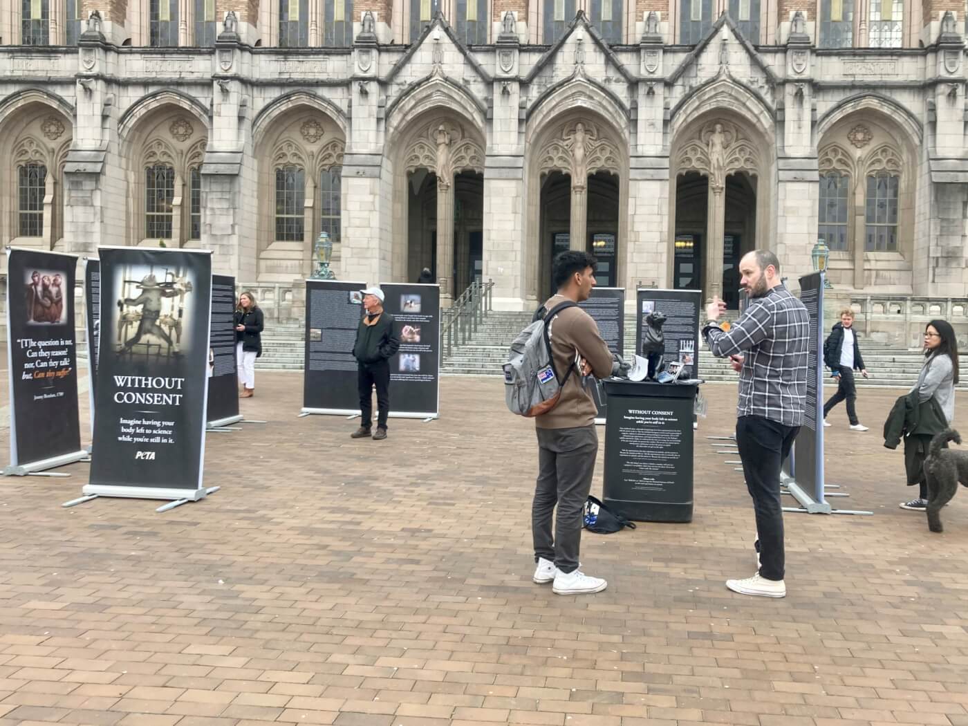 Photo of University of Washington's "Without Consent" exhibit. Photo is taken from outside a building. Various people are looking at the banner displays.