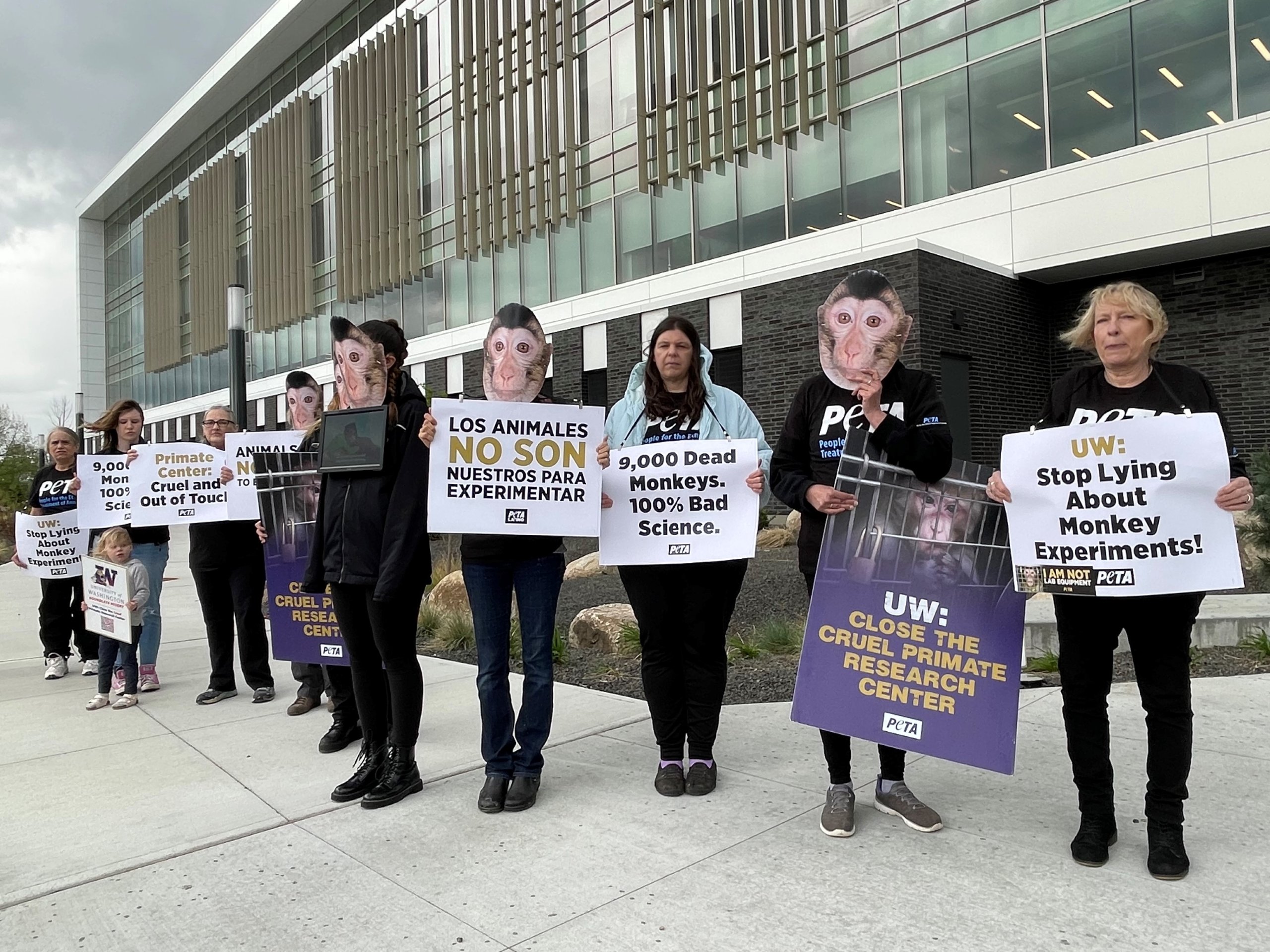 Ten demonstrators stand in a line outside of University of Washington’s health sciences center holding signs in both English and Spanish. Sign text reads: "UW: Stop Lying about Monkey Experiments!", "9,000 Dead Monkeys. 100% Bad Science", "Los Animales No Son Nuestros Para Experimentar", and "Primate Center, Cruel and Out of Touch"
