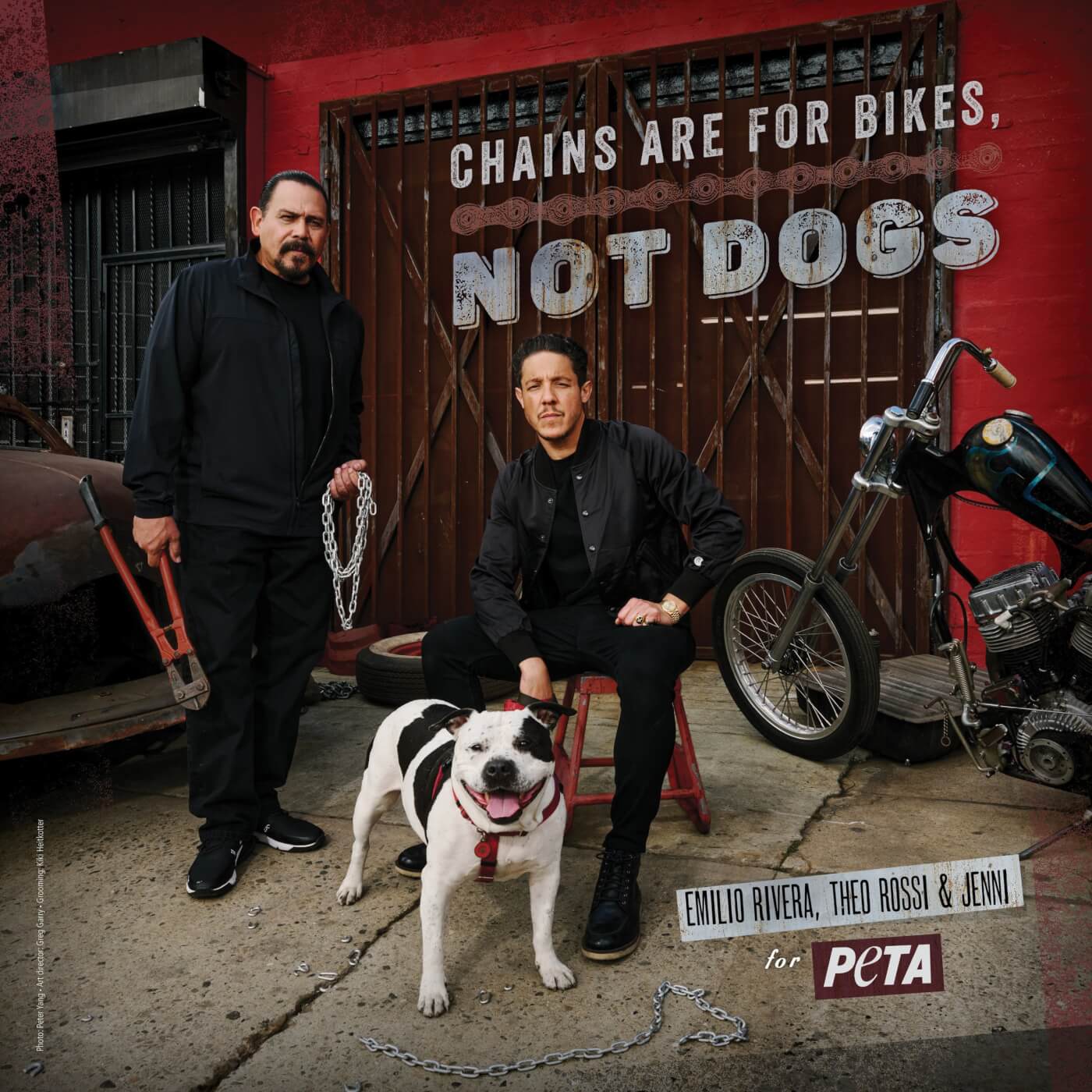 Theo Rossi and Emilio Rivera pose in front of a gate with Jenni, a black and white dog, at their feet. There is a broken chain at her paws. Rivera holds a pair of metal cutters in one hand, and the remainder of the chain in the other.