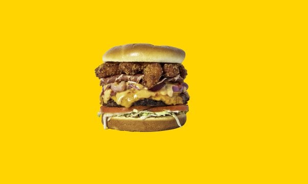 Slutty Vegan Menage a Trois burger with fried vegan shrimp and vegan bacon on top on a yellow background