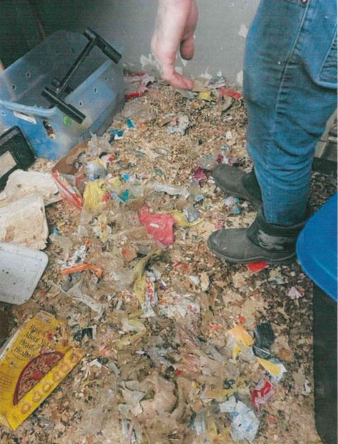 yet another view of a filthy and neglectful reptile store in Plymouth, Pennsylvania, with more unsanitary cages, trash and feces on the floor, and clutter everywhere as a human's legs are visible standing in the mess