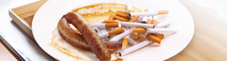 text at top reads "Still serving meat? How about a cigarette, too" with image in the middle of a plate on a hospital food tray with cigarettes and meat sausages. The text below reads "Eating processed meat increases cancer risk. NHS, go vegan."