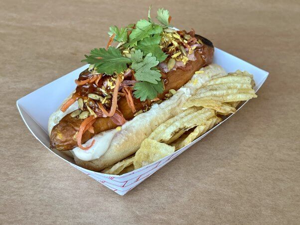 Vegan carrot dog in a bun topped with veggies, cilantro, and sauce served with chips