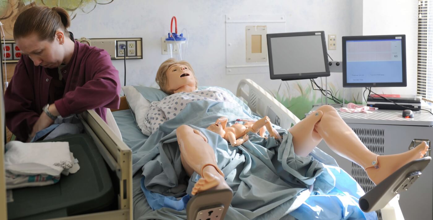 a person in scrubs to the left of a hospital bed, which has a rubber human simulator on it, legs spread.