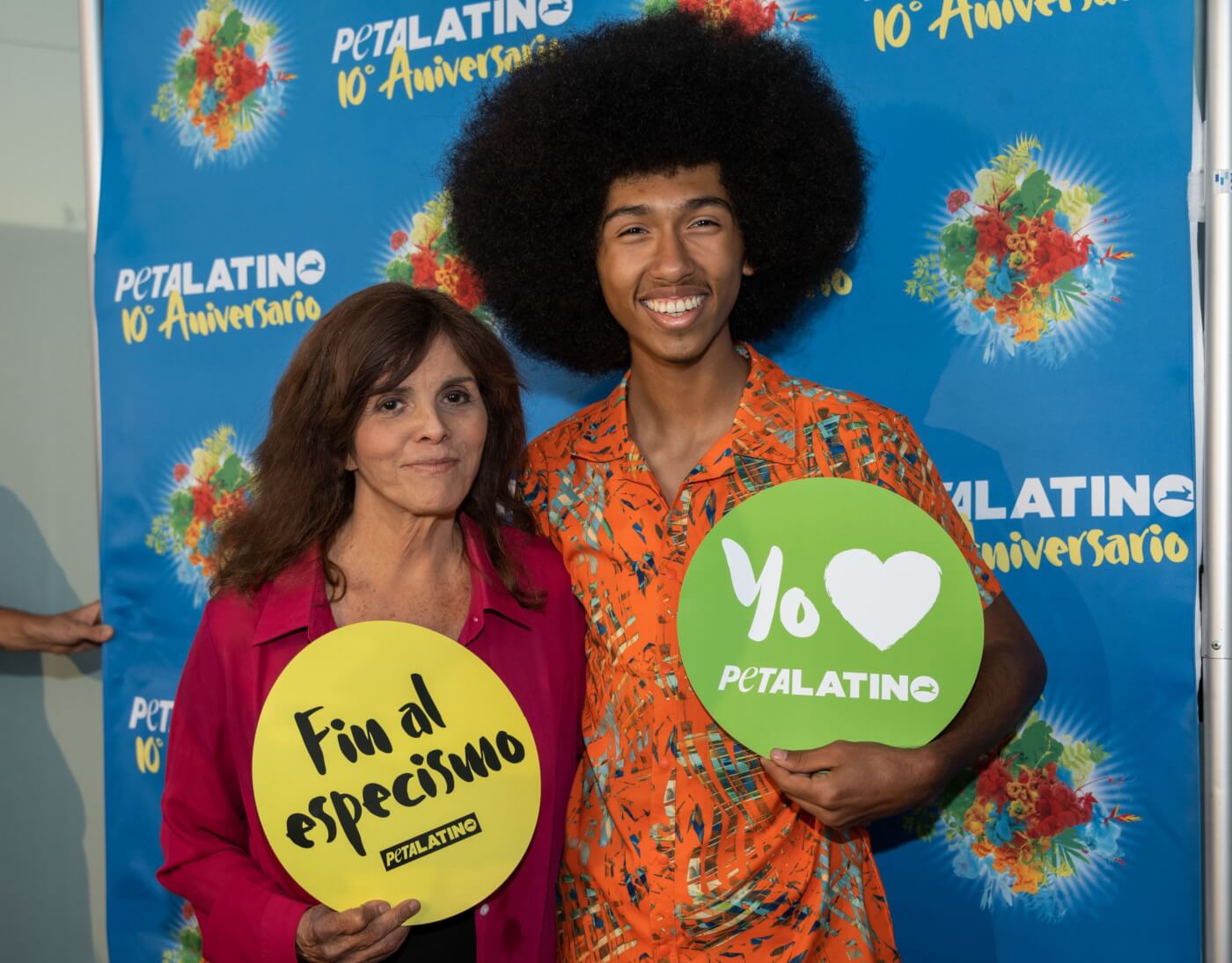 two attendees of the PETA Latino tenth anniversary party, holding round animal advocacy signs in front of the celebratory banner