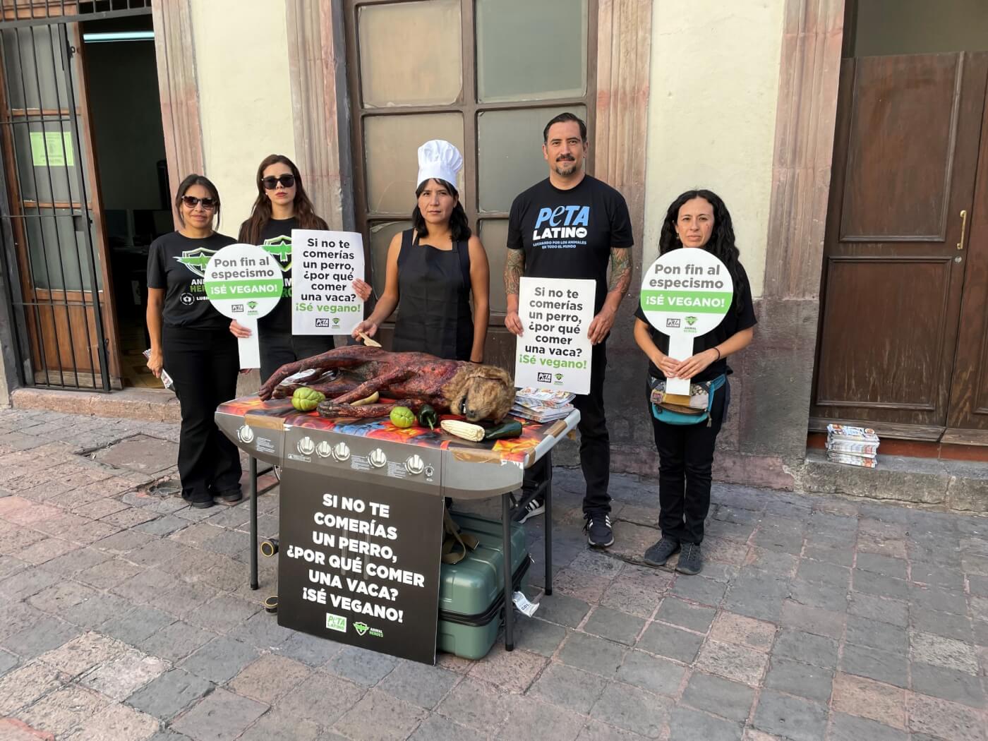 five people in animal heroes and peta latino shirts standing in front of a building. in front of them is a mock grill with a realistic prop dog, which appears to be skinned and charred.