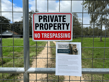 a metal gate with a sign reading "Private property no trespassing". Behind the gate is a field of green grass, and further behind is a building and a treeline.