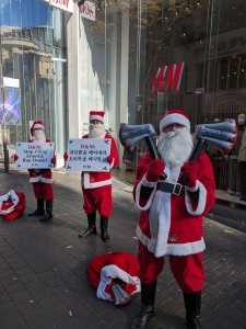 In front of an H&M store. Three people dressed as Santa Claus. Two hold signs, one holds two inflatable bloody axe prop. Signs from left to right: H&M Stop elf ing around, ban down! The next sign is in Hangul and I'm not a translator.