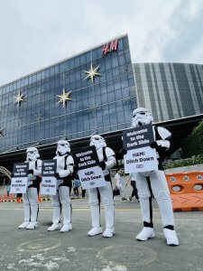 In front of an H&M store. Four people dressed in Storm Trooper costumes from Star Wars in a row holding identical signs. Signs read