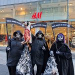 In front of an H&M store. Three people dressed as skeleton reapers, holding sickles that read: H&M: Down is Dead. The center of the group holds up a prop of a bloodied duck, and the two on each side hold jackets with dirty feathers adhered to them.