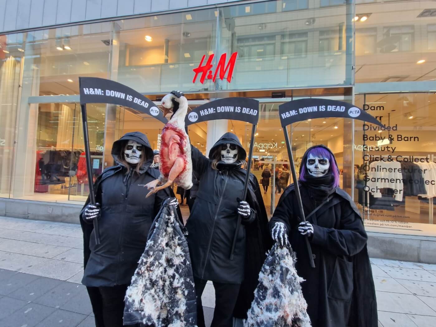 In front of an H&M store. Three people dressed as skeleton reapers, holding sickles that read: H&M: Down is Dead. The center of the group holds up a prop of a bloodied duck, and the two on each side hold jackets with dirty feathers adhered to them.