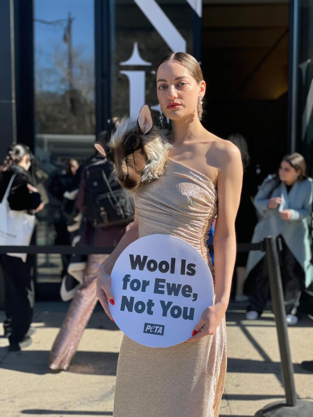 a model in a dress with a fake sheep head on their shoulder. they hold a sign reading "wool is for ewe (note: spelled as in baby sheep) not you (note: as in the personal pronoun)"