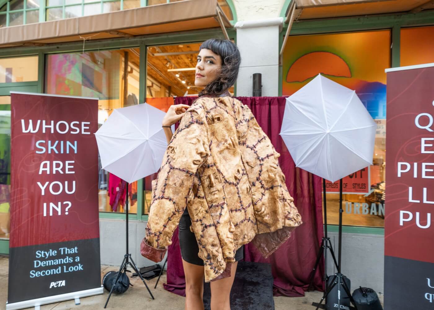 a person poses with a jacket made to look like it's made of human faces. signs in the background read "whose skin are you in?"