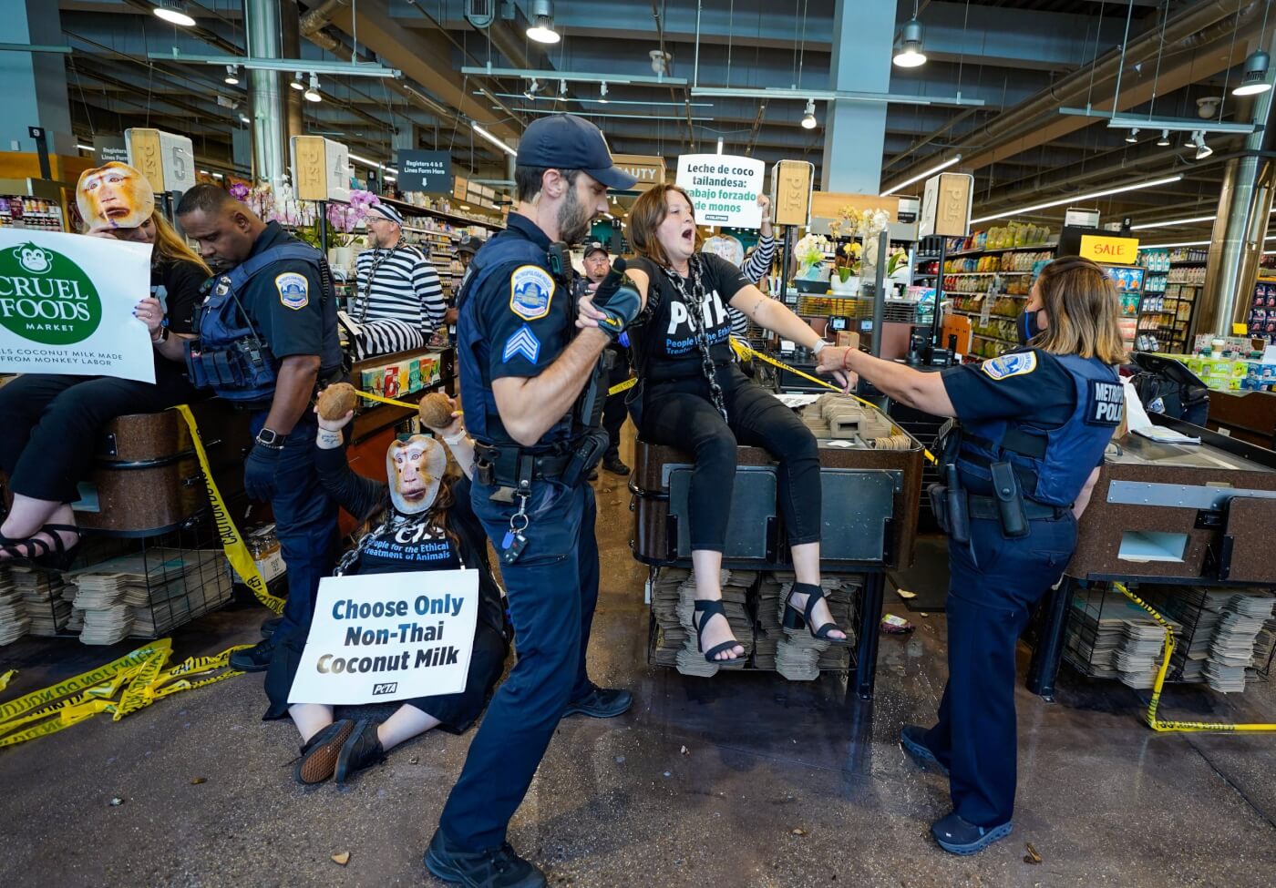 protesters blockading the checkout lanes at whole foods, with monkey masks and signs reading "cruel foods" and "choose only non thai coconut milk". other protestors appear in mock prison jumpsuits. police officers are grabbing protestors.