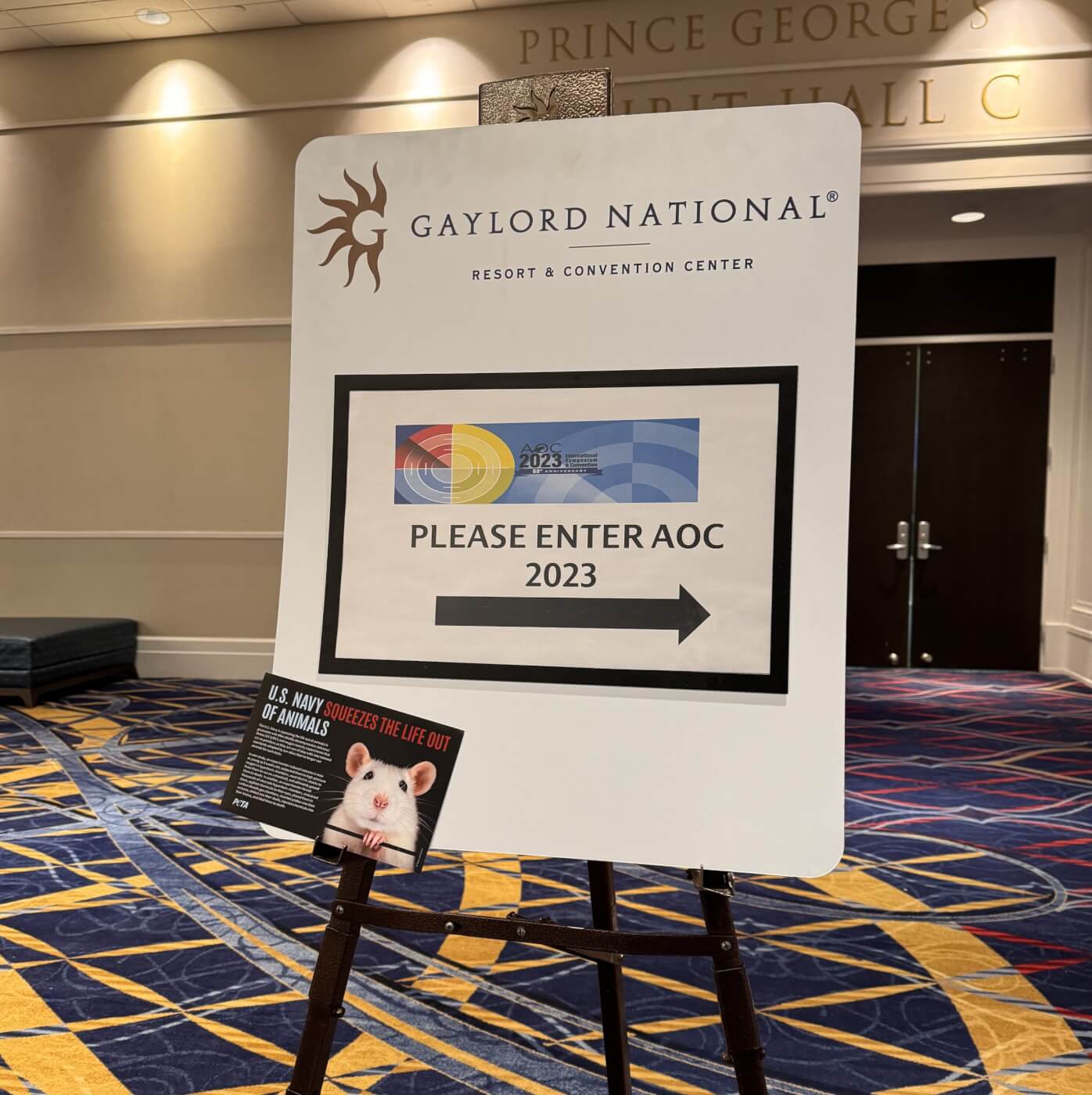 Photo of a stand-up sign for the Gaylord National Resort pointing the way to AOC 2023. There is a flyer taped to the corner from PETA with the top line reading U.S. Navy Squeezes the Life Out of Animals