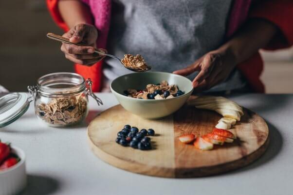 woman preparing a bowl of healthy vegan cereal, with sliced apples, bananas, and blueberries