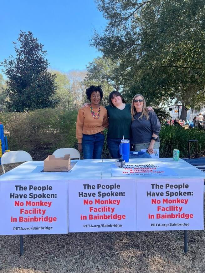 image shows a trio of people standing behind a row of signs attached to a table - text reads: The people have spoken: no monkey facility in Bainbridge