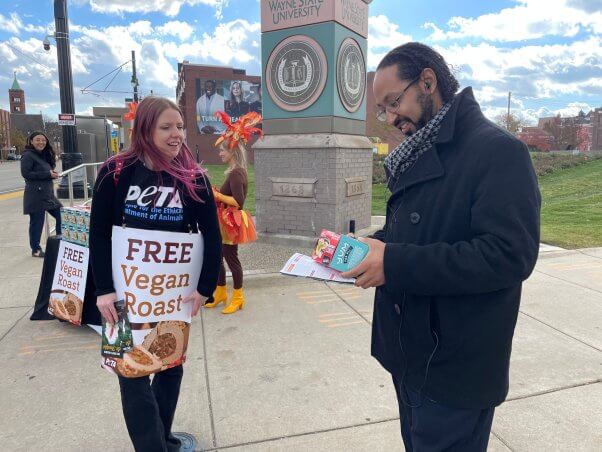 A PETA demonstrator wearing a 'free vegan roast' sandwich sign talks to a passerby