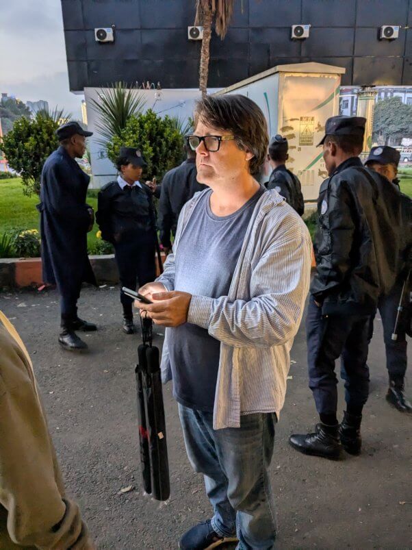 photo of Jason Baker standing, using smartphone phone with uniformed officials behind him in background