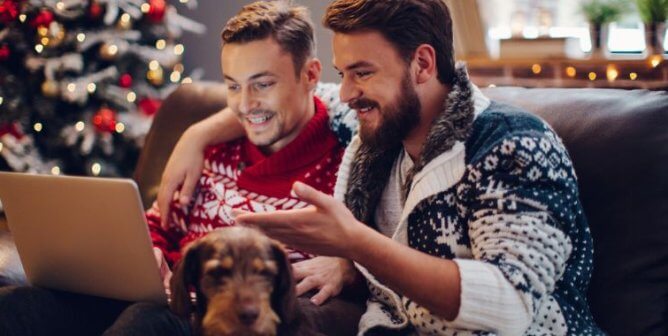 A couple wearing festive sweaters shopping online. There is a dog resting between them