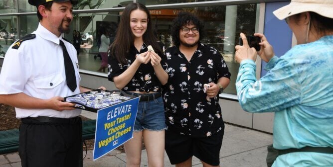 Man dressed in pilot uniform with tray of vegan cheese, two people posing, and someone taking a picture