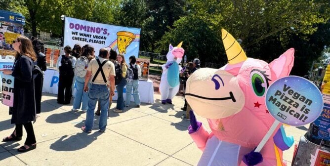 A unicorn mascot holding a 'Vegan Pizza is Magical!' sign