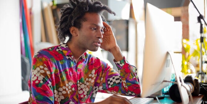 Pensive man wearing colorful shirt, sitting at the desk and using computer in his studio, Photographer at work.