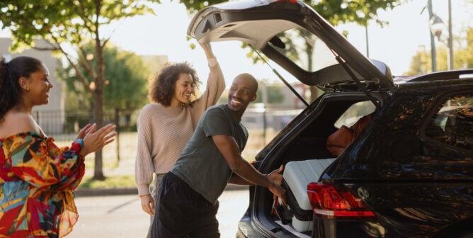 Happy friends packing luggage into car for road trip